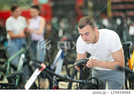 Attentive young guy choosing model seat on high-speed bike in sports shop Attentive young guy choosing model seat on high-speed bike in sports shop 129454688