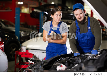 Woman with documents looks at guy mechanic repairing car under hood Woman with documents looks at guy mechanic repairing car under hood 129454710