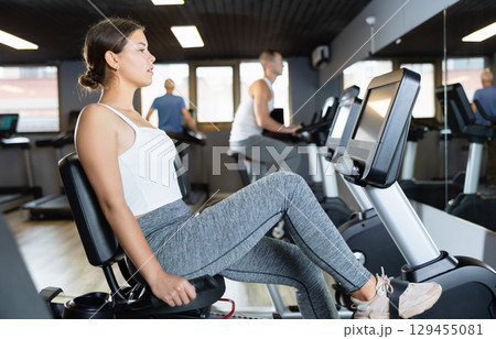 Concentrated young girl working out on stationary bicycle in gym. Healthy active lifestyle concept 129455081