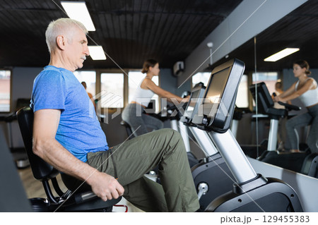 Elderly man, along with other people, is engaged on an bike in gym Elderly man, along with other people, is engaged on an bike in gym 129455383