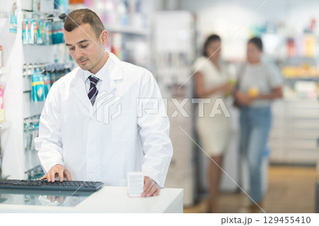 Young male pharmacist standing at the desk using keyboard in chemist's shop 129455410