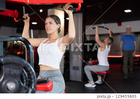 Girl doing strength training in gym, exercising upper body muscles on shoulder press machine Girl doing strength training in gym, exercising upper body muscles on shoulder press machine 129455603