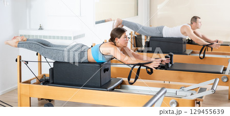 Girl works out on in gym on Pilates simulator. Sports club client on reformer with cable and rope performs exercise to stretch muscles of arms in gym 129455639
