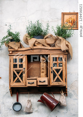 Wooden wall-mounted cupboard decorated with burlap-wrapped herb planters, small bags, vintage cookware, and a rustic painting. Cottagecore decor, pastoral aesthetics, cozy countryside 129456021
