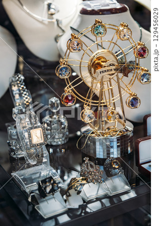 Close-up of a jewelry shop window featuring ornate rings, luxury watches, and a Ferris wheel-themed gem display. Luxury accessories, visual merchandising, gift economy, retail storytelling Close-up of a jewelry shop window featuring ornate rings, luxury watches, and a Ferris wheel-themed gem display. Luxury accessories, visual merchandising, gift economy, retail storytelling 129456029