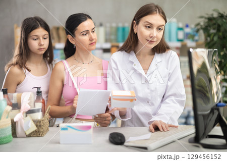 Portrait of young woman pharmacist standing at prescription counter and working on computer, helping mother and daughter select prescription drugs at pharmacy shop 129456152