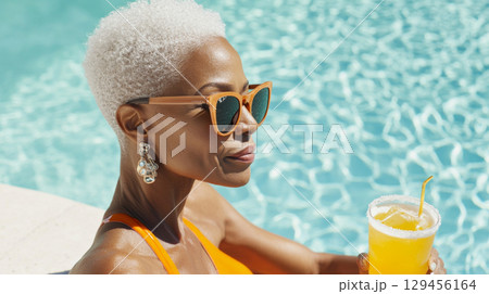 Mature black woman with sunglasses enjoying orange cocktail drink by swimming pool, summer vacation 129456164