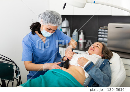 Focused young female cosmetologist in a protective mask makes an elderly woman client a vacuum massage of the abdomen Focused young female cosmetologist in a protective mask makes an elderly woman client a vacuum massage of the abdomen 129456364