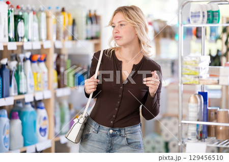 Young woman looking for something on shelf of showcase, reviews assortment in household shop Young woman looking for something on shelf of showcase, reviews assortment in household shop 129456510