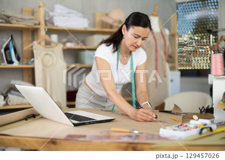 Woman tailor draws dress pattern of product on paper, laptop on table. Woman tailor draws dress pattern of product on paper, laptop on table. 129457026