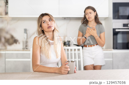 Middle-aged woman sitting at the table while a girl is disputing with her standing behind 129457036