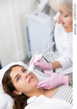 Young woman at appointment with beautician doing ultrasound face cleaning with help of high-frequency ultrasonic waves in cosmetology clinic 129457323