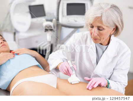 Young woman getting ultrasound cavitation body contouring treatment, anti-cellulite therapy, vacuum massage from old female cosmetologist. Hardware cosmetology for body sculpting 129457547