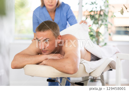 Woman physiotherapist covers man lying on massage table with white terry towel. 129458001