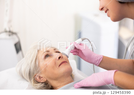 Young female doctor, dermatologist using ultrasound cavitation machine for improvement of skin condition of old female in clinic Young female doctor, dermatologist using ultrasound cavitation machine for improvement of skin condition of old female in clinic 129458390