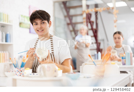 Happy young guy painting on ceramic cup during pottery class 129458402