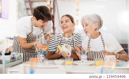 Cheerful older woman with young adult children painting pottery in family workshop 129458406