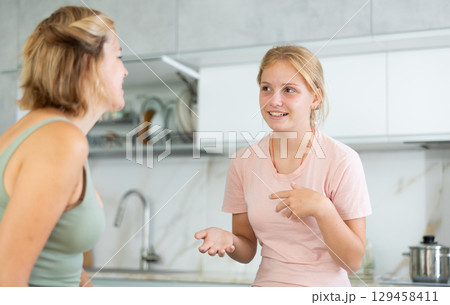 Cheerful teenage daughter talking friendly to mother in kitchen Cheerful teenage daughter talking friendly to mother in kitchen 129458411