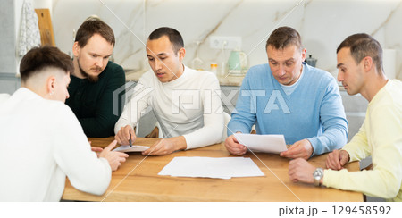 Friends reading financial documents and trying to solve problems together while sitting at table in their kitchen Friends reading financial documents and trying to solve problems together while sitting at table in their kitchen 129458592