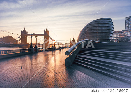 A beautiful morning at Queen's Walk showcases Tower Bridge towering over the River Thames. Soft light casts reflections on the pavement, creating a serene atmosphere for walkers. 129458967