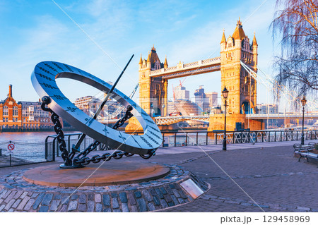 A large sundial stands prominently by the Thames River, with Tower Bridge in the background. The sun casts shadows on the sundial, marking the time while tourists enjoy the view. A large sundial stands prominently by the Thames River, with Tower Bridge in the background. The sun casts shadows on the sundial, marking the time while tourists enjoy the view. 129458969