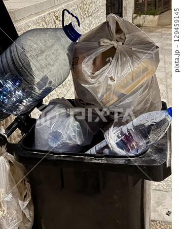 Black outdoor trash bin overflowing with plastic bags and water bottles sits on patterned stone walkway, drawing attention to urban waste and environmental impact, close up 129459145