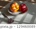Close-up photo of 2 ripe peach (yellow, red) in floral bowl on light table, with spoon and pink flower 129460089