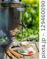 Close-up photo of a silver samovar & teacup on wooden table outdoors, with an ornate urn & small cup (filled with light liquid) nearby. Scattered red candies & checkered saucer. Bright, natural 129460090