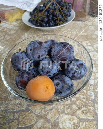Plums and an apricot in a glass bowl in a realistic still life style in purple, orange, and beige colors, representing healthy eating and harvest 129461276