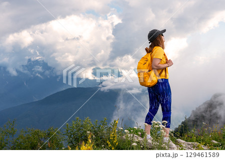 Young woman hiker in yellow shirt and backpack stands on rocky mountain peak, gazing at breathtaking landscape with clouds and distant mountains, embodying adventure and exploration, Active lifestyle Young woman hiker in yellow shirt and backpack stands on rocky mountain peak, gazing at breathtaking landscape with clouds and distant mountains, embodying adventure and exploration, Active lifestyle 129461589