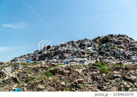 A massive pile of trash dominates the landscape, surrounded by scattered debris and greenery under a bright sky A massive pile of trash dominates the landscape, surrounded by scattered debris and greenery under a bright sky 129462070