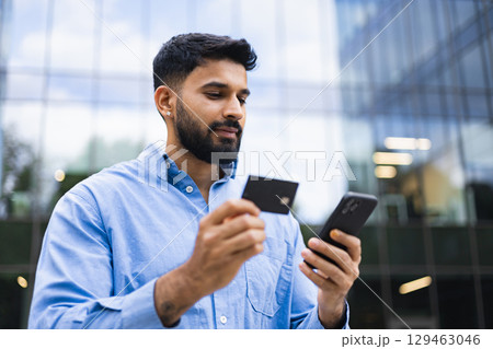 An Indian man in a blue shirt is shown holding a credit card and a smartphone in front of a modern building. 129463046