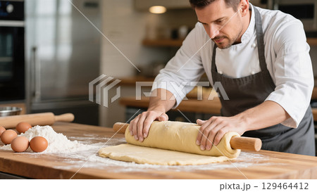 Focused male chef rolling dough with wooden pin on floured table in professional kitchen 129464412