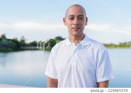 Confident bald man in white polo shirt posing by lake in serene outdoor portrait Confident bald man in white polo shirt posing by lake in serene outdoor portrait 129465570