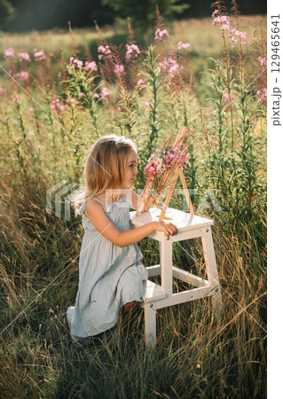 Young girl painting flowers in a meadow surrounded by tall grass during golden hour 129465641