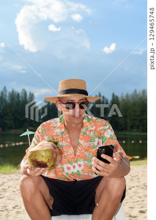 Man tourist sitting on beach using mobile phone and holding coconut water 129465748