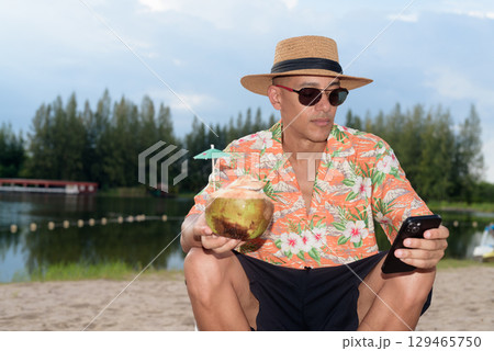 Man tourist sitting on beach using mobile phone and holding coconut water 129465750
