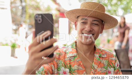 Hispanic man using phone taking selfie at outdoor cafe wearing Panama hat and short sleeve shirt 129465870