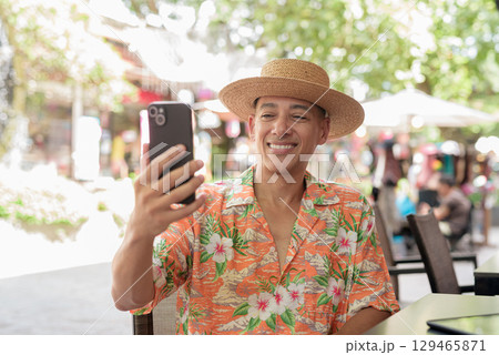 Hispanic man using phone taking selfie at outdoor cafe wearing Panama hat and short sleeve shirt Hispanic man using phone taking selfie at outdoor cafe wearing Panama hat and short sleeve shirt 129465871