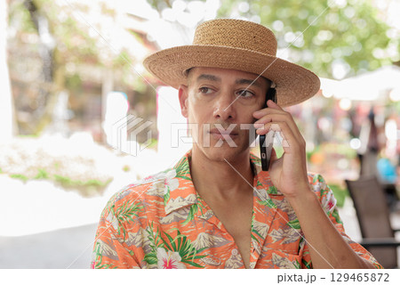 Hispanic man talking on phone at outdoor cafe wearing Panama hat and short sleeve shirt 129465872