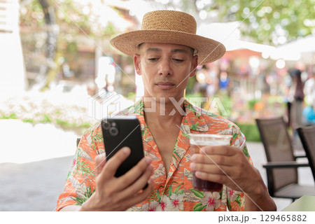 Hispanic man holding iced coffee and using phone at cafe wearing Panama hat and Hawaiian shirt 129465882
