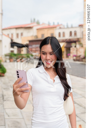 Asian woman using phone in white polo shirt on European style city street during summer Asian woman using phone in white polo shirt on European style city street during summer 129466107
