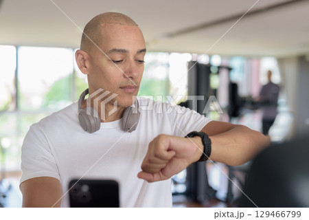 Latin man cycling on stationary exercise bike in gym while using mobile phone and smartwatch 129466799