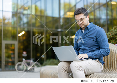 Professional individual focused on work using a laptop outside a contemporary glass building, depicting productivity in a serene outdoor setting. 129467176
