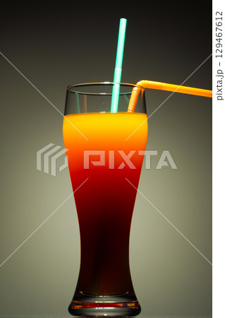 A tall glass containing a layered beverage, likely a Tequila Sunrise cocktail, against a dark, muted background 129467612
