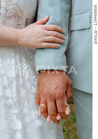 Close-up image of a bride and groom gently holding hands, showcasing their wedding rings. The bride wears a lace dress, and both express a sense of unity and commitment. 129469806