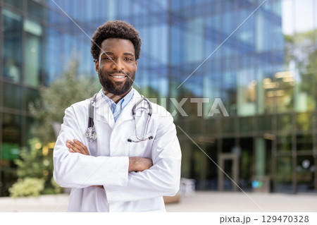 Portrait of a smiling male physician in a white coat, standing confidently outside a modern medical facility and symbolizing health, care, and professionalism within contemporary urban setting. Portrait of a smiling male physician in a white coat, standing confidently outside a modern medical facility and symbolizing health, care, and professionalism within contemporary urban setting. 129470328