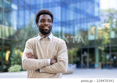 A confident individual is standing outside with arms crossed, near a modern glass structure. 129470341