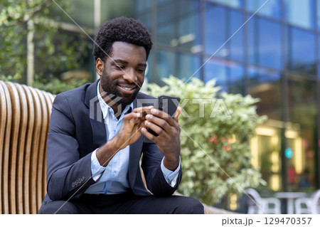 Confident young professional using a phone outdoors at a contemporary business location, showcasing technology and connectivity in a beautiful and productive work setting. 129470357