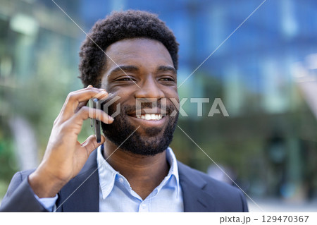 Businessman in a suit smiling while speaking on a mobile phone outdoors, portraying confidence and communication in a professional setting. Businessman in a suit smiling while speaking on a mobile phone outdoors, portraying confidence and communication in a professional setting. 129470367